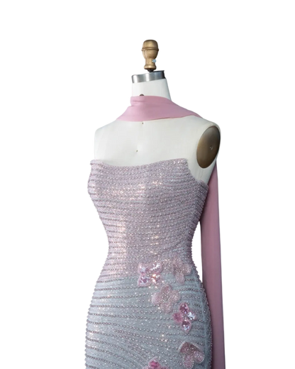 A strapless evening gown displayed on a dress form mannequin against a pale blue background. The dress is covered in shimmering silver sequins with pink floral applique and a matching pink draped scarf.